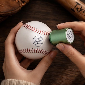 May include: A white baseball with red stitching featuring a circular black ink stamp of a baseball and a signature. A cylindrical green ink stamp tool is pressed against the surface of the ball, displaying the same circular logo on its top end.