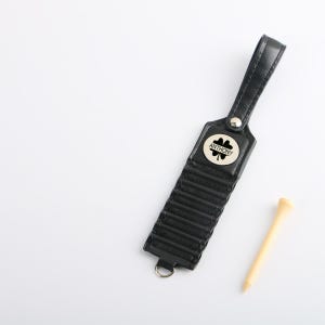 May include: Black leather golf tee holder with a silver-colored button and a small metal emblem with the name "ANTHONY" and a four-leaf clover. A wooden golf tee is next to the holder. The holder has a loop for attachment.