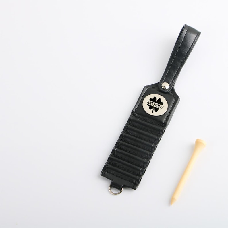 May include: Black leather golf tee holder with a silver-colored button and a small metal emblem with the name "ANTHONY" and a four-leaf clover. A wooden golf tee is next to the holder. The holder has a loop for attachment.