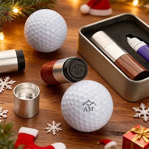 May include: A collection of golf-themed gifts, including personalized golf balls with mountain and initial designs, a wooden and metal golf ball marker, and a metal container with a golf ball marker and pen. The items are arranged on a wooden surface with Christmas decorations.
