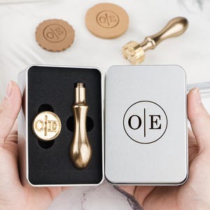 Puede incluir: Un juego de sellos de lacre de color dorado con las iniciales "O|E" dentro de una lata plateada. El juego incluye un sello, un mango y dos sellos de cera. La tapa de la lata también presenta las iniciales "O|E".