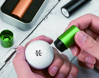 May include: A white golf ball with the black text KIEVEN printed on it, being stamped by a green cylindrical ink marker. Additional ink markers in copper and black colors are visible in a metal storage tin nearby.
