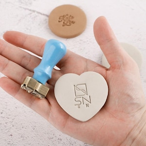 May include: A light blue handled wax stamp with a brass stamp head, next to a heart-shaped stamp with the letters "SN" and a round stamp with the text "Kate Lin". The stamps are light beige.