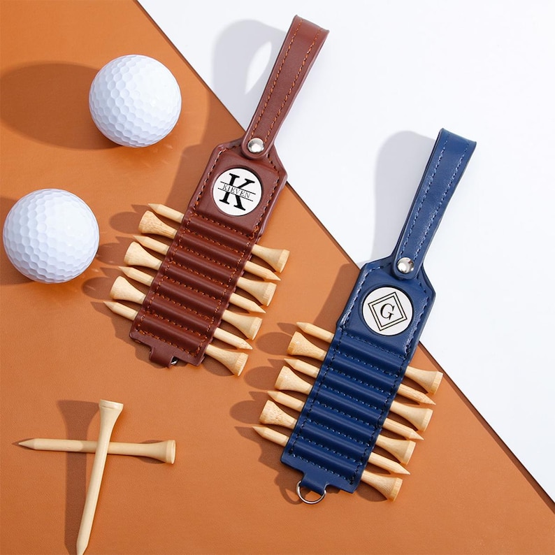 May include: Two golf tee holders, one brown and one blue, each with a leather strap and a circular emblem with a letter. Each holder contains wooden golf tees. Two white golf balls and a few loose tees are also visible.