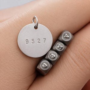 Puede incluir: Un colgante circular de metal plateado con los números 9527 estampados, acompañado de cuatro pequeñas herramientas de estampado de metal cuadradas con caracteres grabados, dispuestas en una fila vertical junto al colgante.
