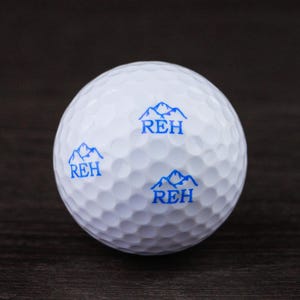 Peut inclure: Une balle de golf blanche avec une surface alvéolée. La balle présente trois motifs bleus de montagnes et de texte qui se lisent "REH". La balle de golf est conçue pour un usage récréatif.