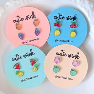 Op de afbeelding: Vier sets fruit-thema oorbellen op roze en blauwe kaartjes. Elk set bevat twee oorbellen met verschillende vruchten. De kaartjes hebben de tekst "cutie wink" en de social media handle "@cutiewinkco".