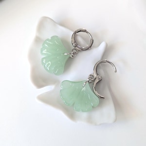 May include: A pair of earrings featuring light green, translucent ginkgo leaf-shaped charms suspended from silver-toned metal hoop clasps. The leaf charms have a smooth, molded surface with subtle ridges radiating from the base to the edges.