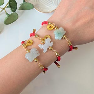 May include: Three red cord bracelets featuring carved light green and white jade-like animal charms. Each bracelet is accented with small gold-toned metal beads and a circular gold-toned charm embossed with a square centre hole design.