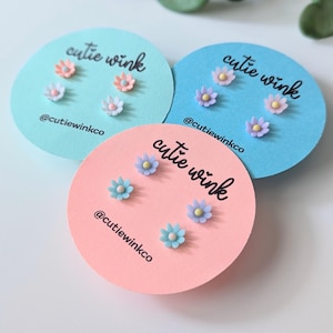 Peut inclure: Trois ensembles de boucles d'oreilles à fleurs sur des cartes circulaires roses et bleues. Chaque carte porte le texte "cutie wink" et le nom de profil sur les réseaux sociaux "@cutiewinkco". Les boucles d'oreilles sont roses, bleues et violettes.