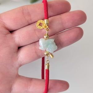 May include: A red cord bracelet featuring a central carved pale green jade-like rabbit charm, flanked by two small gold-toned metal beads. A small circular gold-toned metal charm with a square cutout is attached near the top of the cord.