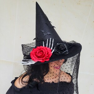 May include: A black pointed witch hat featuring a wide brim adorned with a large red rose, a smaller black rose, and a white skeleton hand. The hat is decorated with black glittery bat cutouts and a sheer black polka dot veil draped over the brim.