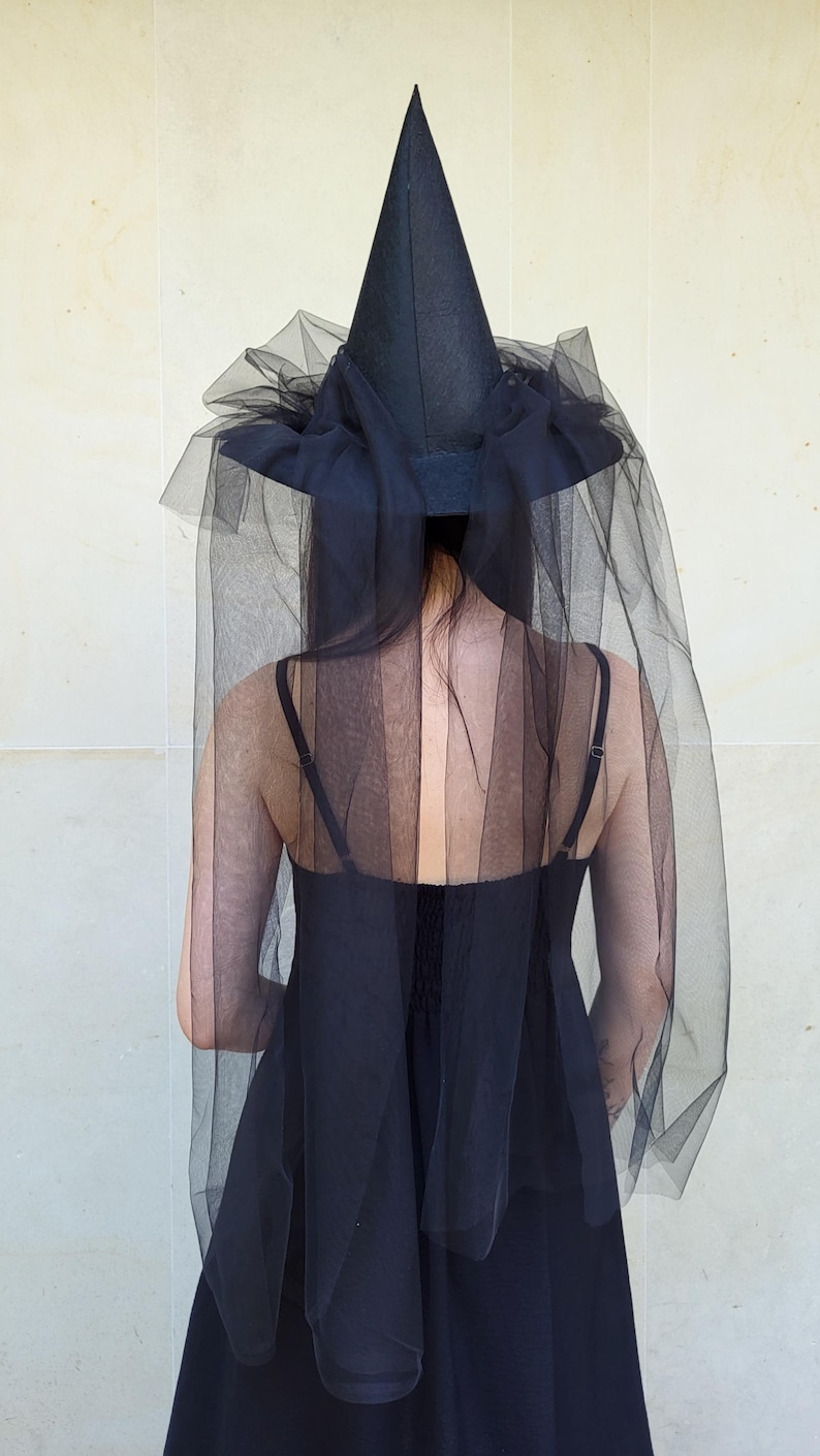 May include: Black witch hat with a tall, pointed crown and a wide brim. The hat is adorned with a black tulle veil that drapes down the back. The hat is part of a Halloween costume.