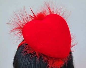 May include: A red heart-shaped headpiece with a velvet texture and feathery accents. The headpiece is worn on black hair, creating a striking contrast. The feathers are a vibrant red, adding to the overall romantic aesthetic.