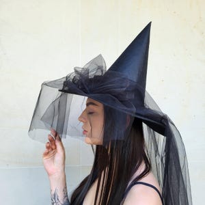 May include: A black witch hat with a tall, pointed crown and a wide brim. The hat is adorned with a large bow and a sheer veil that drapes down. The hat is made of a dark, possibly satin-like material, and is suitable for Halloween or costume events.