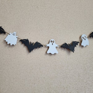 May include: A Halloween garland featuring alternating white ghost and black bat cutouts strung together on a thin brown twine. The ghosts have simple facial expressions with cut-out eyes and mouths, while the bats have detailed wing shapes.