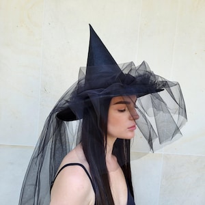 May include: A black witch hat with a tall, pointed crown and a wide brim. The brim is adorned with layers of black tulle, and a long veil of the same material drapes down. This hat is perfect for Halloween or costume parties.