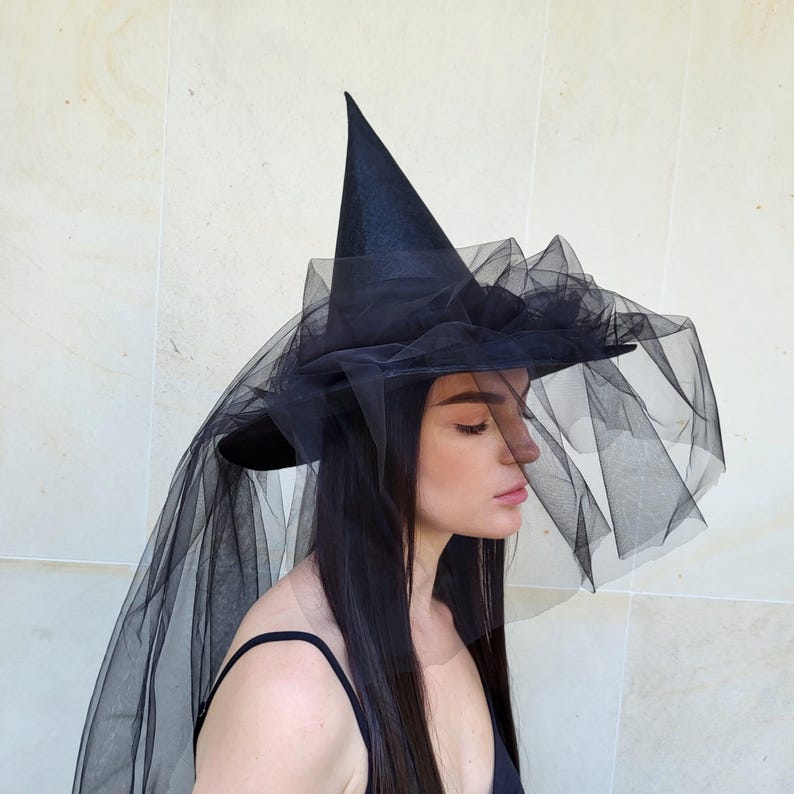 May include: A black witch hat with a tall, pointed crown and a wide brim. The brim is adorned with layers of black tulle, and a long veil of the same material drapes down. This hat is perfect for Halloween or costume parties.