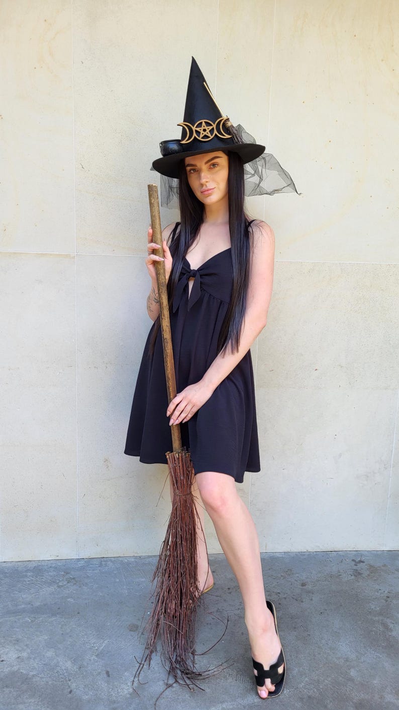 May include: A black witch hat with gold accents and a sheer veil, paired with a black dress and a broom. The hat features a crescent moon and star design. The broom is made of wooden handle and twigs.