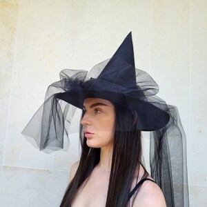 May include: A black witch hat with a tall, pointed crown and a wide brim. The hat is adorned with layers of black tulle, creating a flowing, ethereal effect. The hat is a costume accessory.