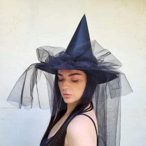 May include: A black witch hat with a tall, pointed crown and a wide brim. The hat is adorned with layers of black tulle, creating a dramatic and elegant effect. The tulle drapes down, adding to the hat's mysterious allure. Perfect for Halloween or costume parties.