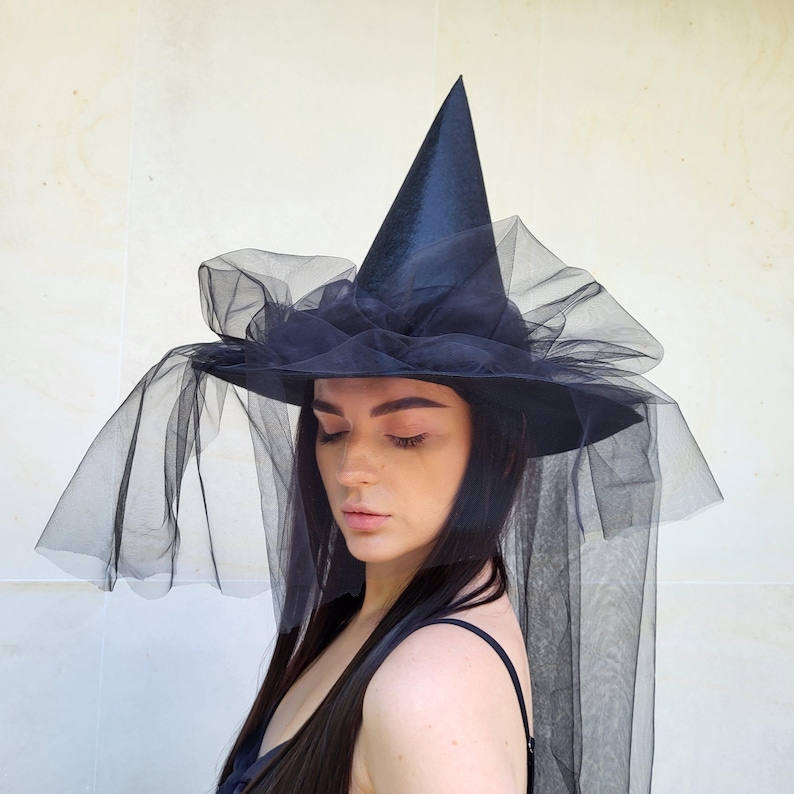 May include: A black witch hat with a tall, pointed crown and a wide brim. The hat is adorned with layers of black tulle, creating a dramatic and elegant effect. The tulle drapes down, adding to the hat's mysterious allure. Perfect for Halloween or costume parties.