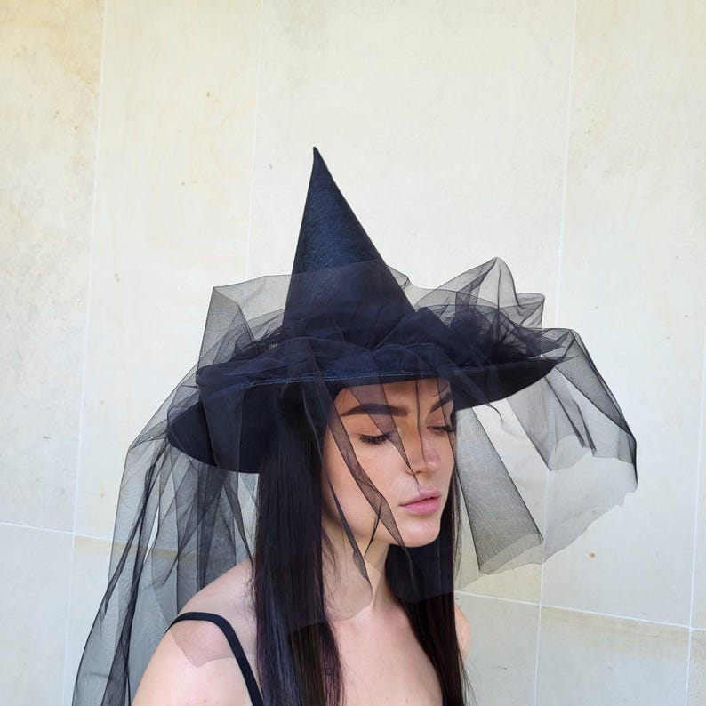 May include: A black witch hat with a wide brim and a tall, pointed crown. The hat is adorned with a flowing, sheer black veil. The hat is a costume accessory, perfect for Halloween or themed events.