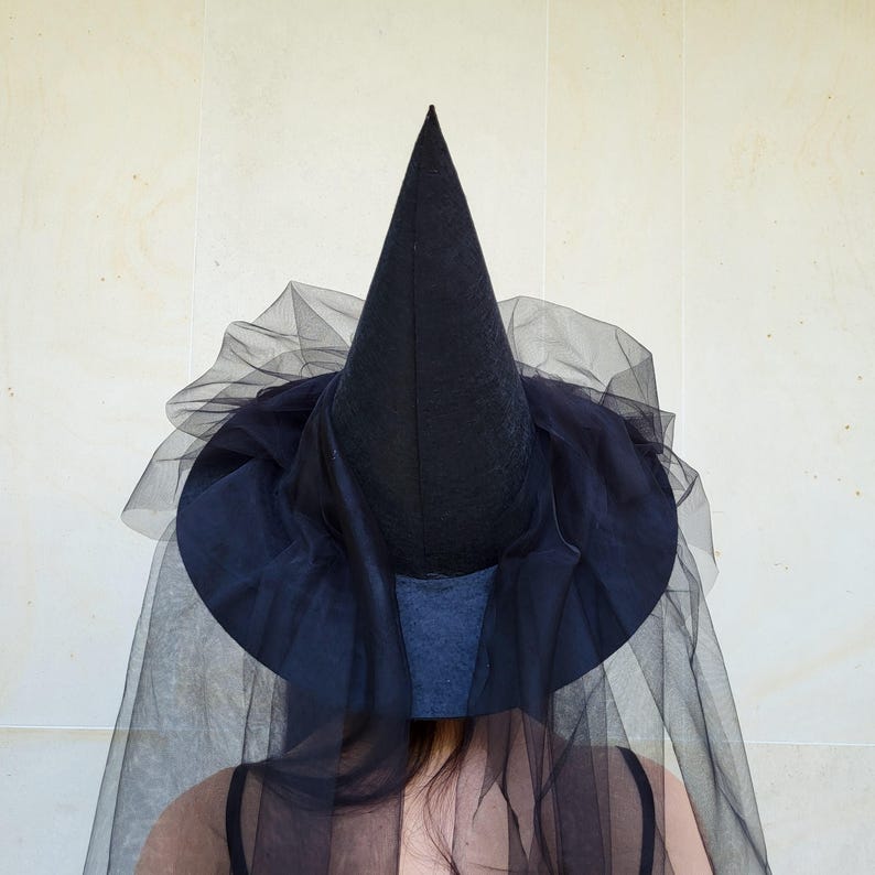 May include: A black witch hat with a tall, pointed crown and a wide brim. The hat is adorned with layers of black tulle, creating a flowing, ethereal effect. The hat is a costume accessory.