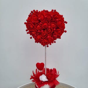 May include: A decorative floral arrangement. A heart-shaped topiary made of red roses and berries sits on a white stem. The base is decorated with red feathers, a small red heart, and a white rose.