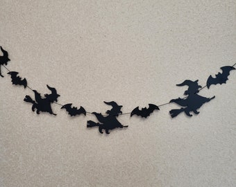 May include: A black decorative garland featuring silhouettes of witches riding broomsticks and flying bats strung together on a thin cord. The cut-out shapes are arranged in a repeating pattern, creating a horizontal display of dark figures.