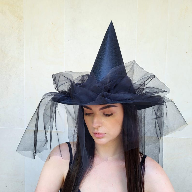 May include: A black witch hat with a tall, pointed crown and a wide brim. The brim is adorned with layers of black tulle, and a veil drapes down. The hat is a costume accessory, perfect for Halloween or themed events.