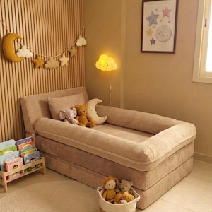 May include: A beige corduroy toddler bed with a low profile, surrounded by a soft, neutral-toned nursery. The room features a moon and star garland, a cloud-shaped lamp, and a framed picture with celestial designs. A basket of stuffed animals sits nearby.