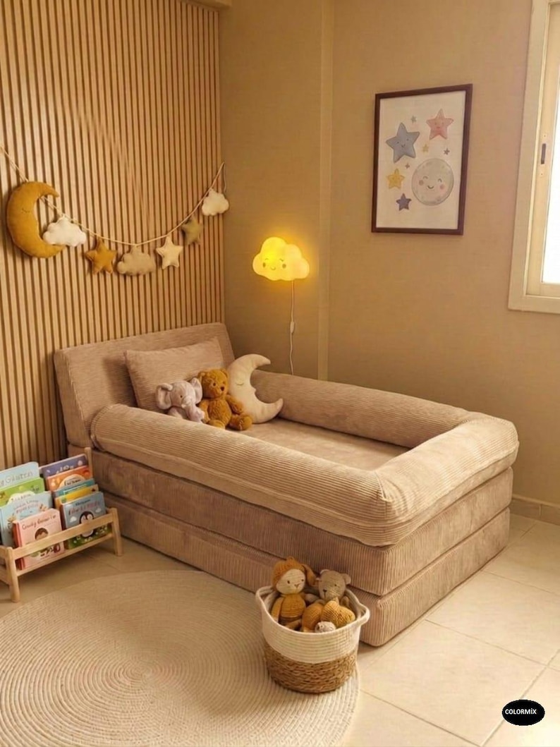 May include: A beige corduroy toddler bed with a low profile, surrounded by a soft, neutral-toned nursery. The room features a moon and star garland, a cloud-shaped lamp, and a framed picture with celestial designs. A basket of stuffed animals sits nearby.