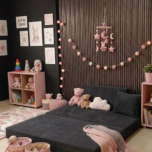 Puede incluir: Una habitaci&oacute;n infantil con un sof&aacute; cama convertible gris oscuro, estanter&iacute;as rosas y decoraci&oacute;n mural. La habitaci&oacute;n est&aacute; decorada con peluches, un m&oacute;vil y una manta rosa y gris. El texto "ELIF'IN ODASI" es visible.