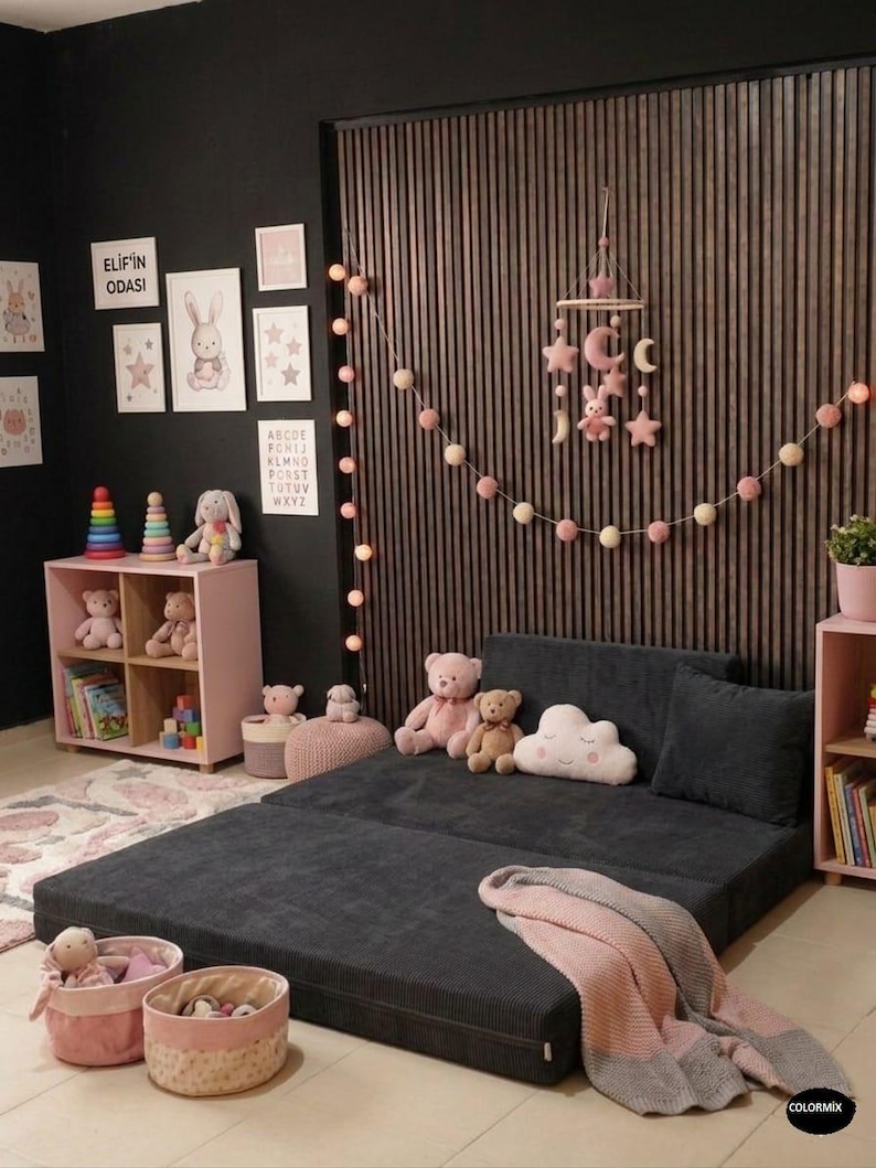 Puede incluir: Una habitaci&oacute;n infantil con un sof&aacute; cama convertible gris oscuro, estanter&iacute;as rosas y decoraci&oacute;n mural. La habitaci&oacute;n est&aacute; decorada con peluches, un m&oacute;vil y una manta rosa y gris. El texto "ELIF'IN ODASI" es visible.