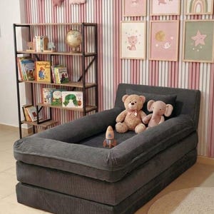 Puede incluir: Una cama infantil de pana gris oscuro con un borde elevado, apilada en dos capas. Dos peluches y un cohete de juguete est&aacute;n dentro. El fondo incluye una estanter&iacute;a, arte enmarcado y una pared a rayas rosas y blancas.