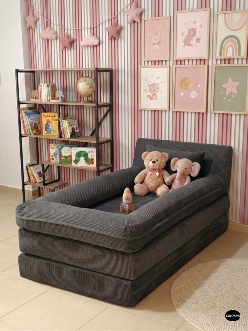 Puede incluir: Una cama infantil de pana gris oscuro con un borde elevado, apilada en dos capas. Dos peluches y un cohete de juguete est&aacute;n dentro. El fondo incluye una estanter&iacute;a, arte enmarcado y una pared a rayas rosas y blancas.