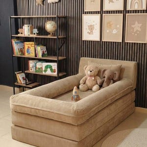 May include: A beige corduroy toddler bed with a raised edge, featuring a teddy bear, an elephant plush toy, and a rocket ship toy. The bed is set against a wall with framed nursery art and a decorative shelf with books and a globe.