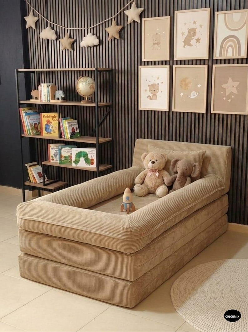 May include: A beige corduroy toddler bed with a raised edge, featuring a teddy bear, an elephant plush toy, and a rocket ship toy. The bed is set against a wall with framed nursery art and a decorative shelf with books and a globe.