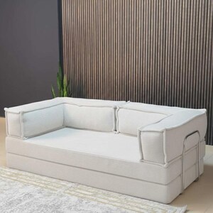 Pu&ograve; includere: Un moderno divano in velluto a coste bianco con un profilo basso. Il divano ha una forma rettangolare con spessi braccioli e schienali imbottiti. Poggia su un tappeto chiaro con un motivo geometrico. Lo sfondo presenta una parete a doghe di legno.