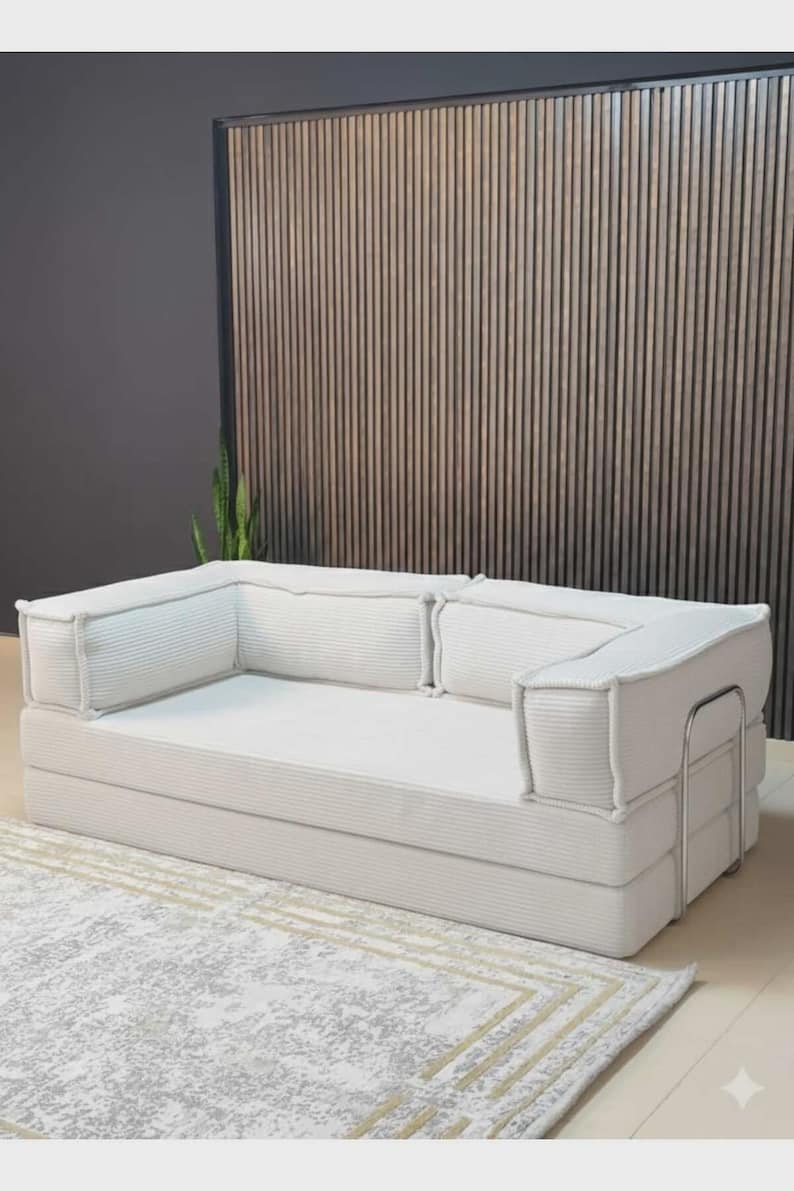 Pu&ograve; includere: Un moderno divano in velluto a coste bianco con un profilo basso. Il divano ha una forma rettangolare con spessi braccioli e schienali imbottiti. Poggia su un tappeto chiaro con un motivo geometrico. Lo sfondo presenta una parete a doghe di legno.