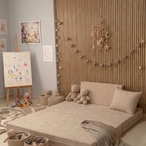 May include: A beige folding floor mat with a matching pillow and blanket. The room features a wooden slatted wall, a drawing easel, and several stuffed teddy bears and toys, creating a cozy children's play area.