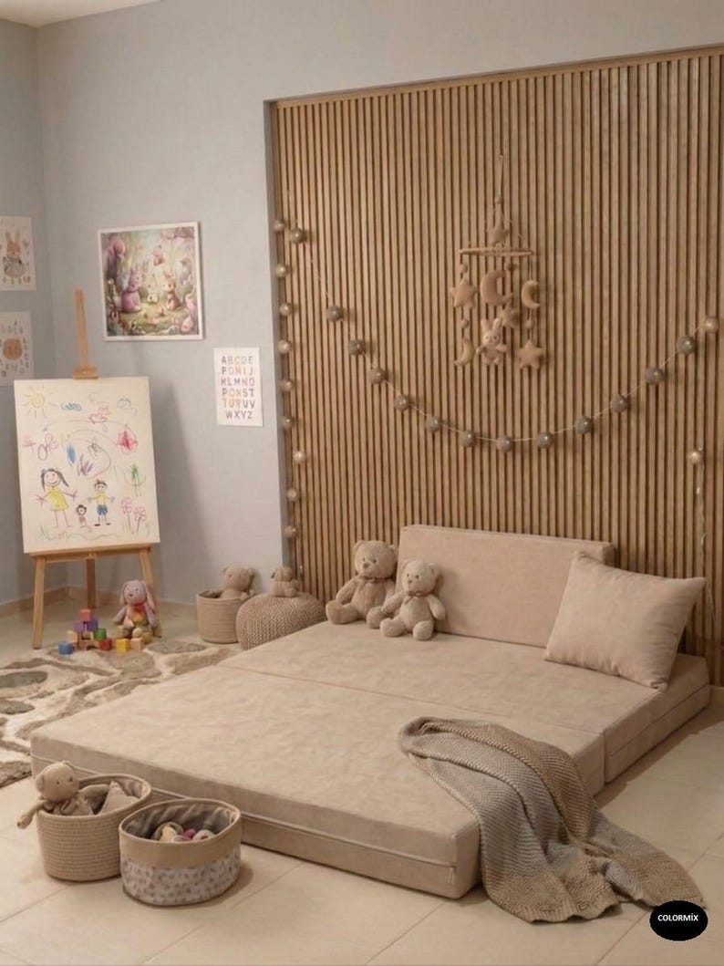 May include: A beige folding floor mat with a matching pillow and blanket. The room features a wooden slatted wall, a drawing easel, and several stuffed teddy bears and toys, creating a cozy children's play area.