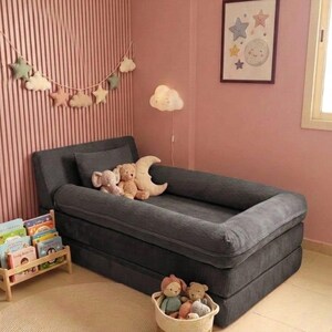 Puede incluir: Una cama para ni&ntilde;os peque&ntilde;os de pana gris con borde elevado, en una habitaci&oacute;n rosa. La cama est&aacute; adornada con animales de peluche. Tambi&eacute;n se ven una alfombra redonda, una cesta de juguetes y una peque&ntilde;a estanter&iacute;a.
