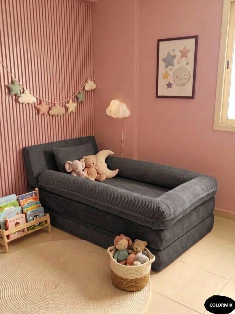 Puede incluir: Una cama para ni&ntilde;os peque&ntilde;os de pana gris con borde elevado, en una habitaci&oacute;n rosa. La cama est&aacute; adornada con animales de peluche. Tambi&eacute;n se ven una alfombra redonda, una cesta de juguetes y una peque&ntilde;a estanter&iacute;a.