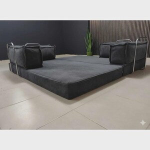 Peut inclure: Un canap&eacute;-lit modulaire gris fonc&eacute; avec une texture en velours c&ocirc;tel&eacute;. Le canap&eacute; a un profil bas et comprend deux coussins carr&eacute;s &agrave; chaque extr&eacute;mit&eacute;, soutenus par des cadres m&eacute;talliques argent&eacute;s. Le lit est pos&eacute; sur un sol carrel&eacute;.