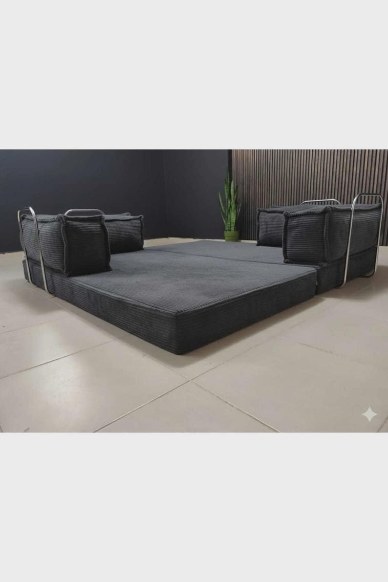 Peut inclure: Un canap&eacute;-lit modulaire gris fonc&eacute; avec une texture en velours c&ocirc;tel&eacute;. Le canap&eacute; a un profil bas et comprend deux coussins carr&eacute;s &agrave; chaque extr&eacute;mit&eacute;, soutenus par des cadres m&eacute;talliques argent&eacute;s. Le lit est pos&eacute; sur un sol carrel&eacute;.