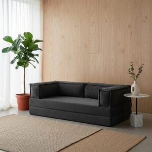 Peut inclure: Un canap&eacute; modulaire gris fonc&eacute; avec un tissu textur&eacute;, des accoudoirs carr&eacute;s et des coussins de dossier. Le canap&eacute; est pos&eacute; sur un tapis marron clair, avec une plante en pot et une petite table d'appoint.