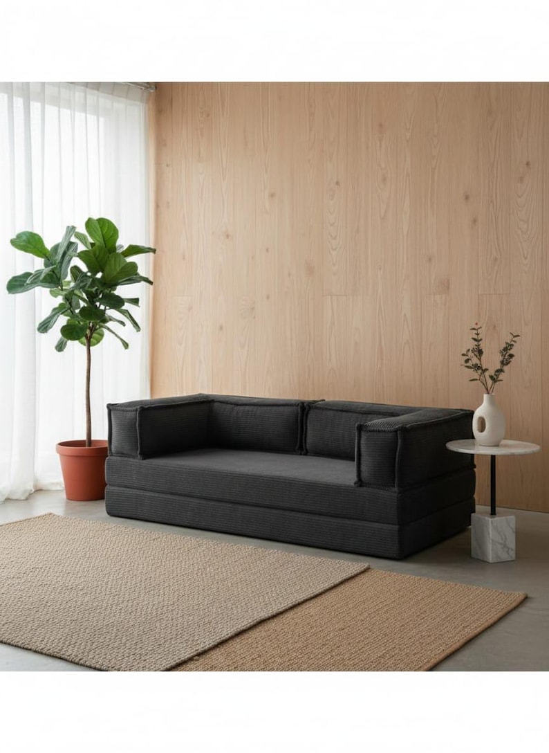 Peut inclure: Un canap&eacute; modulaire gris fonc&eacute; avec un tissu textur&eacute;, des accoudoirs carr&eacute;s et des coussins de dossier. Le canap&eacute; est pos&eacute; sur un tapis marron clair, avec une plante en pot et une petite table d'appoint.