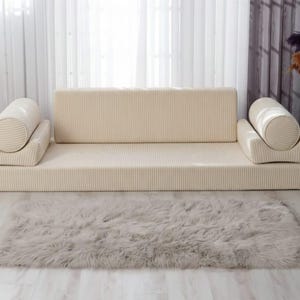 Op de afbeelding: Een beige modulaire bank met een ribfluwelen textuur, bestaande uit een rechthoekige basis, rugleuning en twee cilindrische kussens. Een zacht, grijs rechthoekig vloerkleed ligt voor de bank. De kamer heeft een neutraal kleurenpalet.